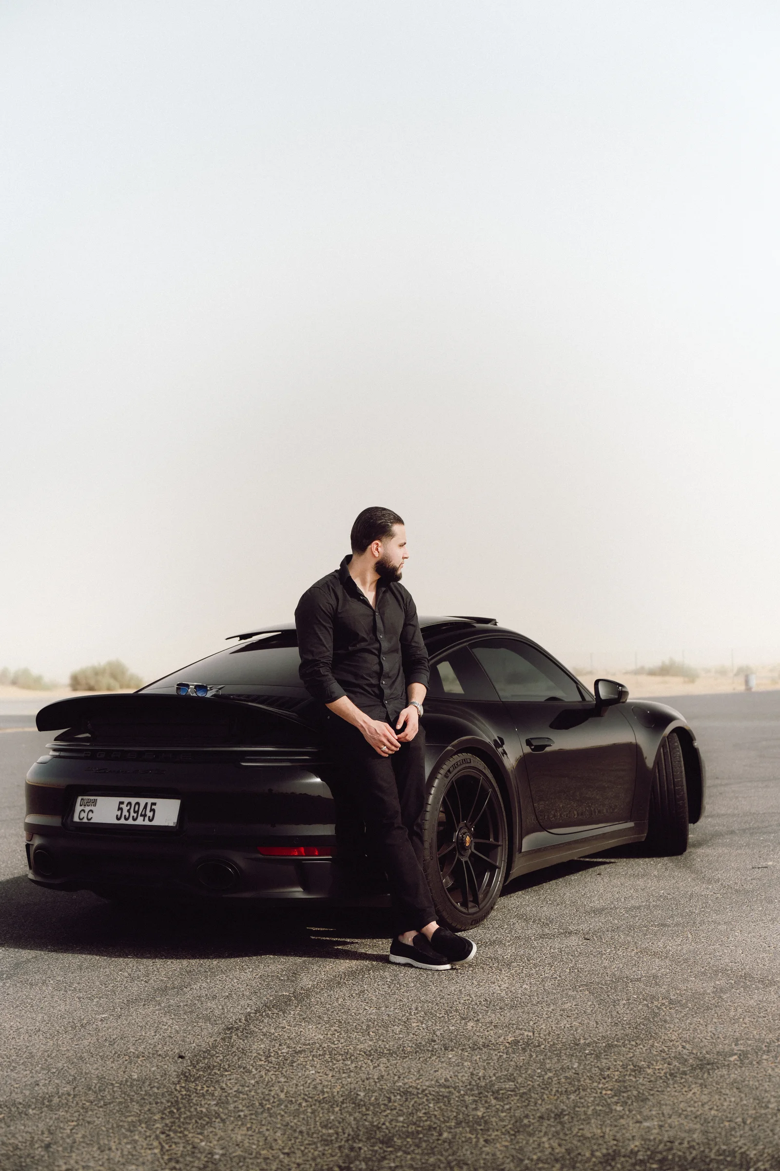 Zack Lalouti leaning against the rear of a black Porsche beside a desert road in Dubai.