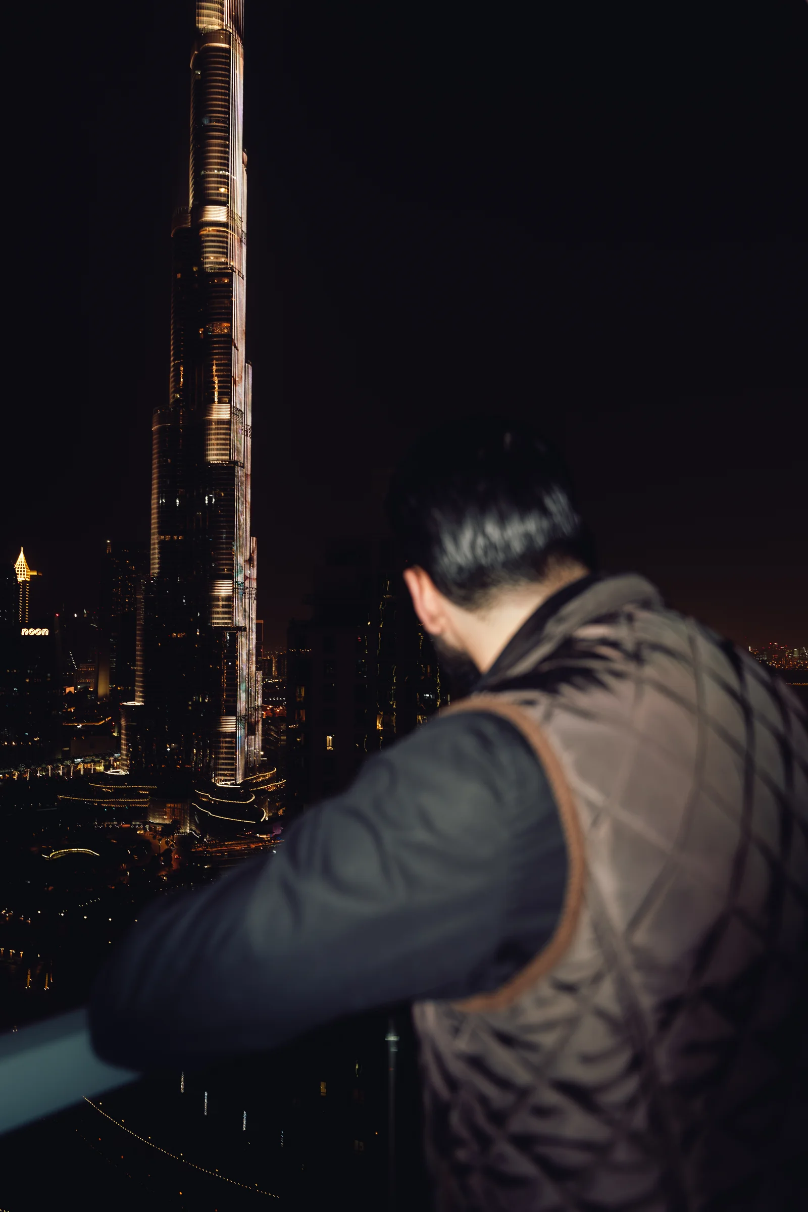 Zack Lalouti leaning on a balcony railing and looking toward the illuminated Burj Khalifa in Dubai at night.