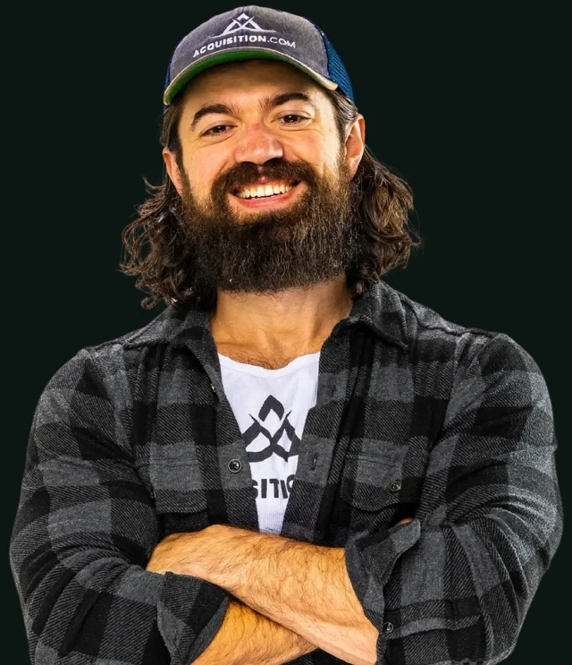 Portrait of Alex Hormozi smiling with his arms folded, wearing an Acquisition.com cap and a dark plaid shirt.